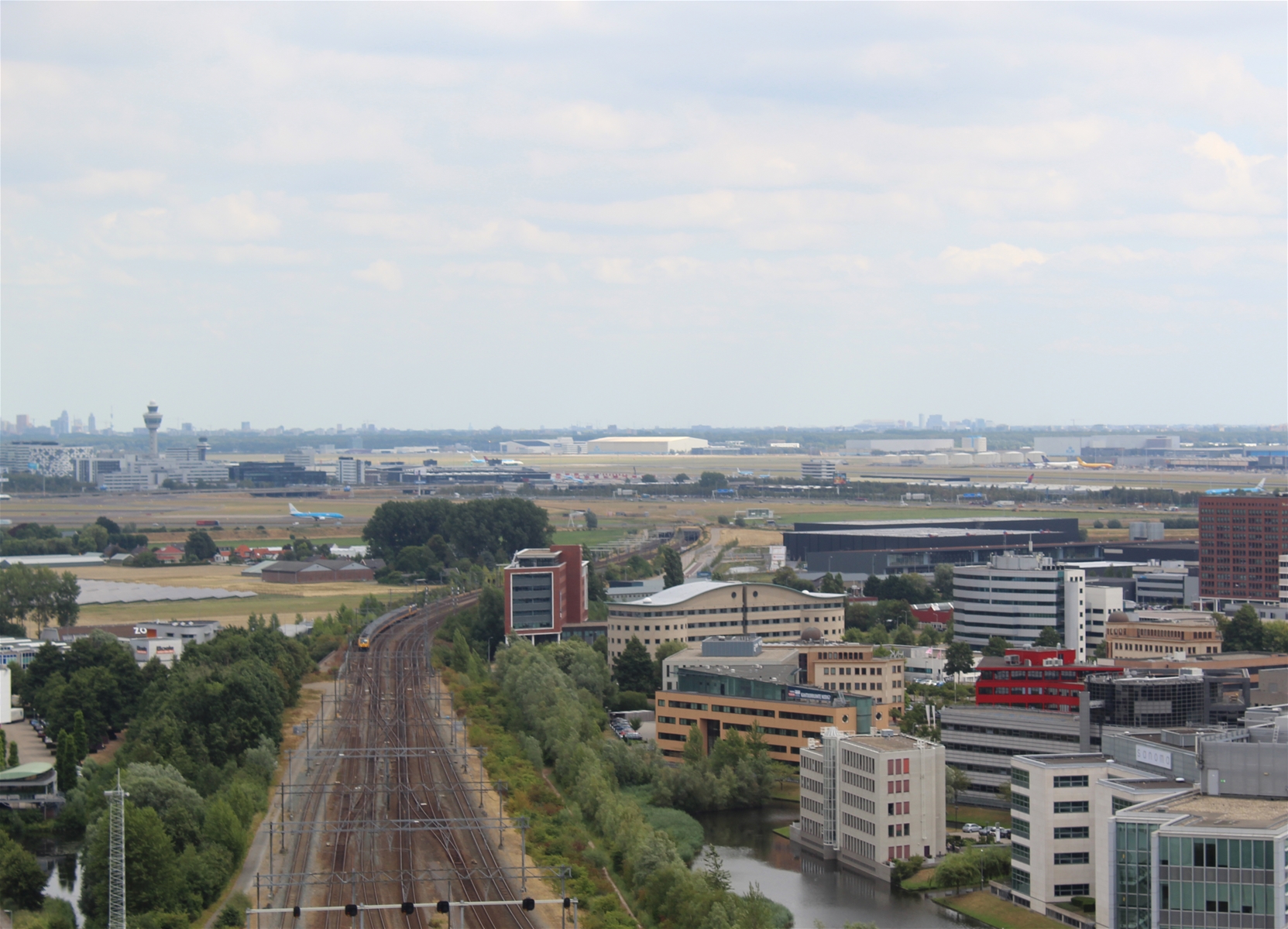 Zicht op Schiphol vanuit een hogere verdieping van het kantoor van de Inspectie Leefomgeving en Transport in Hoofddorp. Op de voorgrond is de spoorlijn Leiden-Schiphol zichtbaar en zijn meerdere kantoorgebouwen zichtbaar van het kantorenpark dat aan Schiphol grenst.
